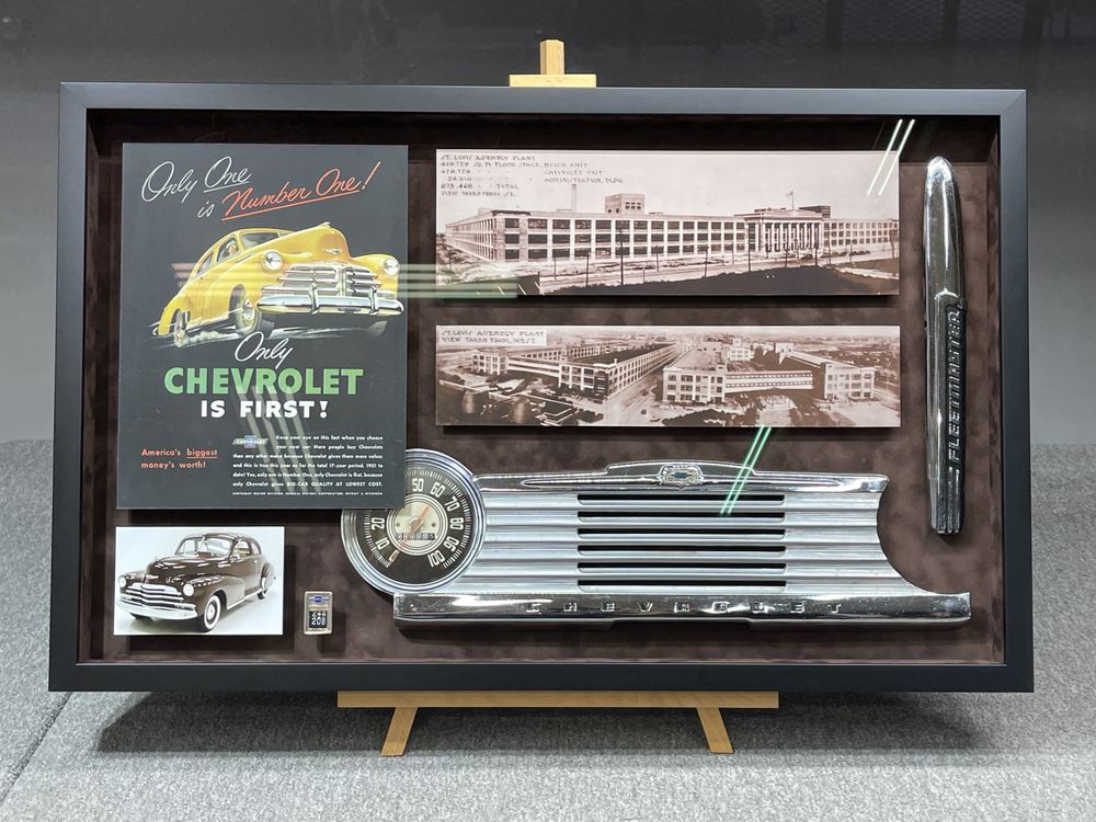 Vintage Chevrolet grille, speedometer, hood ornament, photographs, and advertisement in a custom automotive shadowbox built by Villa